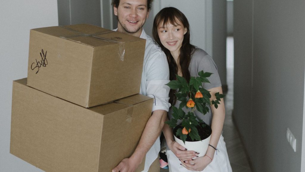 Couple carrying moving boxes and a houseplant into a new home